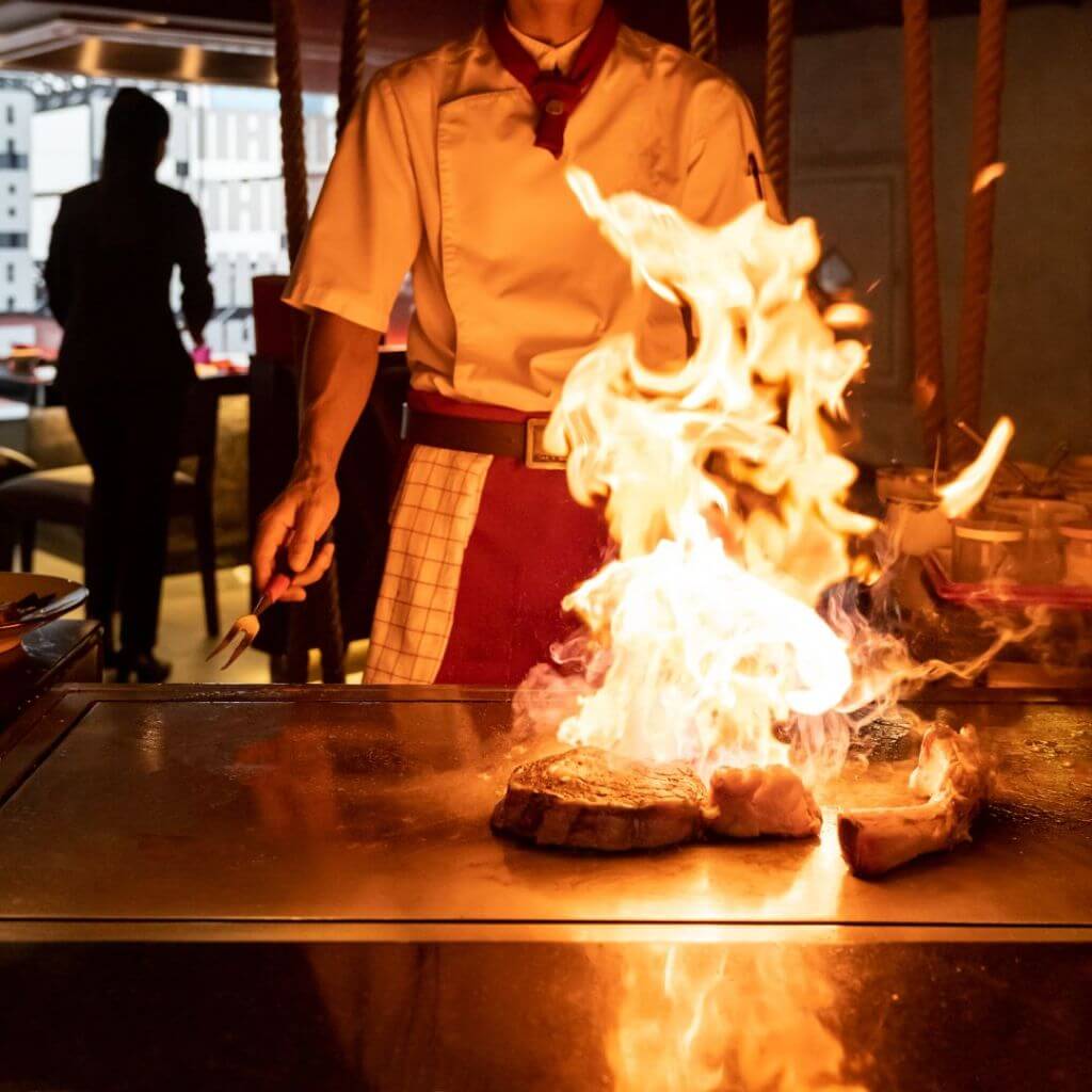 A chef in a restaurant cooking on an open flame grill with fire rising up.