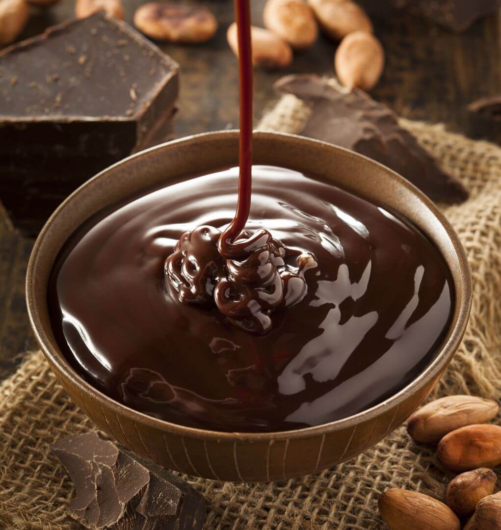 Cocoa syrup drizzling down into a large bowl full of chocolate syrup, surrounded by cocoa beans and chocolate bark