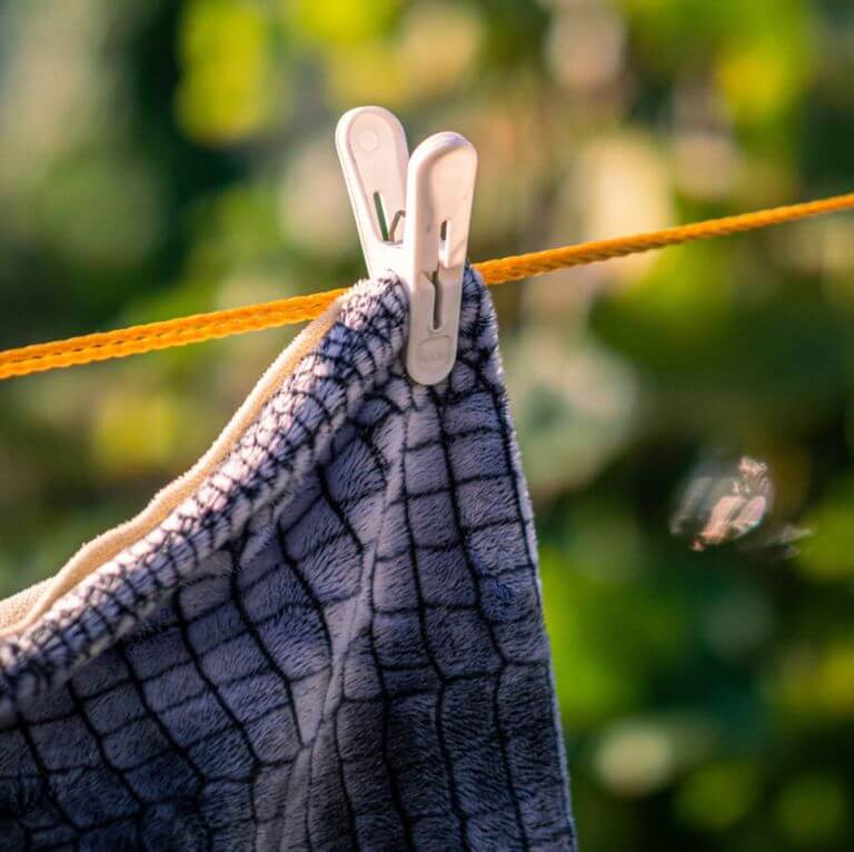 A close-up of a grid-patterned fabric hanging on an orange clothesline outdoors, secured with two white clothespins against a backdrop of green foliage