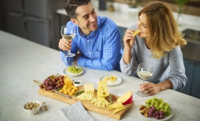 Two persons looking at each other sitting at a table with a platter full of diverse cheeses.