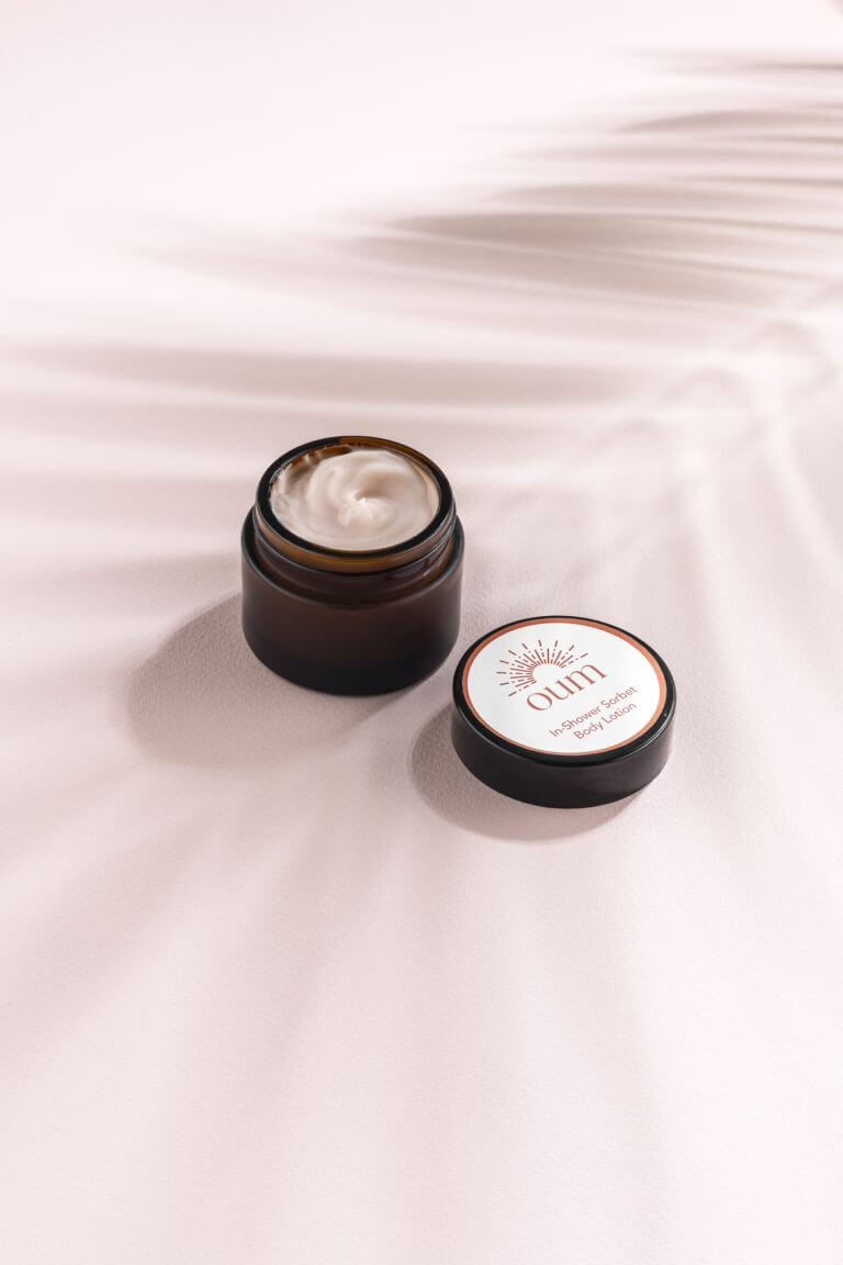 A small open jar of body lotion with a brown container and white cream inside, placed on a light pink surface. The lid is off and lies next to the jar, displaying the brand name