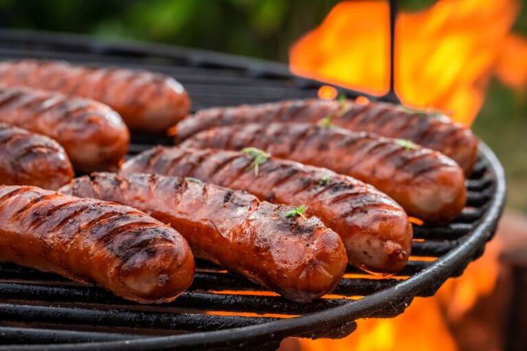 Sausages in a row being grilled on an outdoor grill. Fire rising in the background