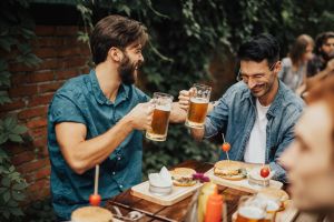 Young men sitting outside, doing cheers and drinking beers. Sandwiches and food items on table around them