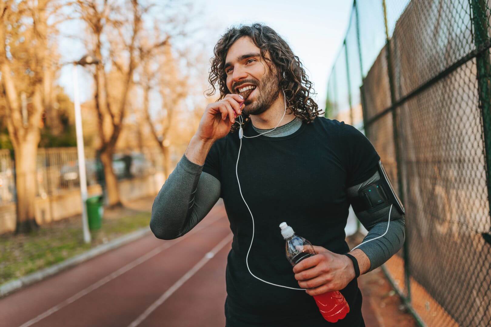 Man working out outside and pausing running to eat a high-protein bar perfect for GLP-1 consumers. He's also holding a bottled beverage