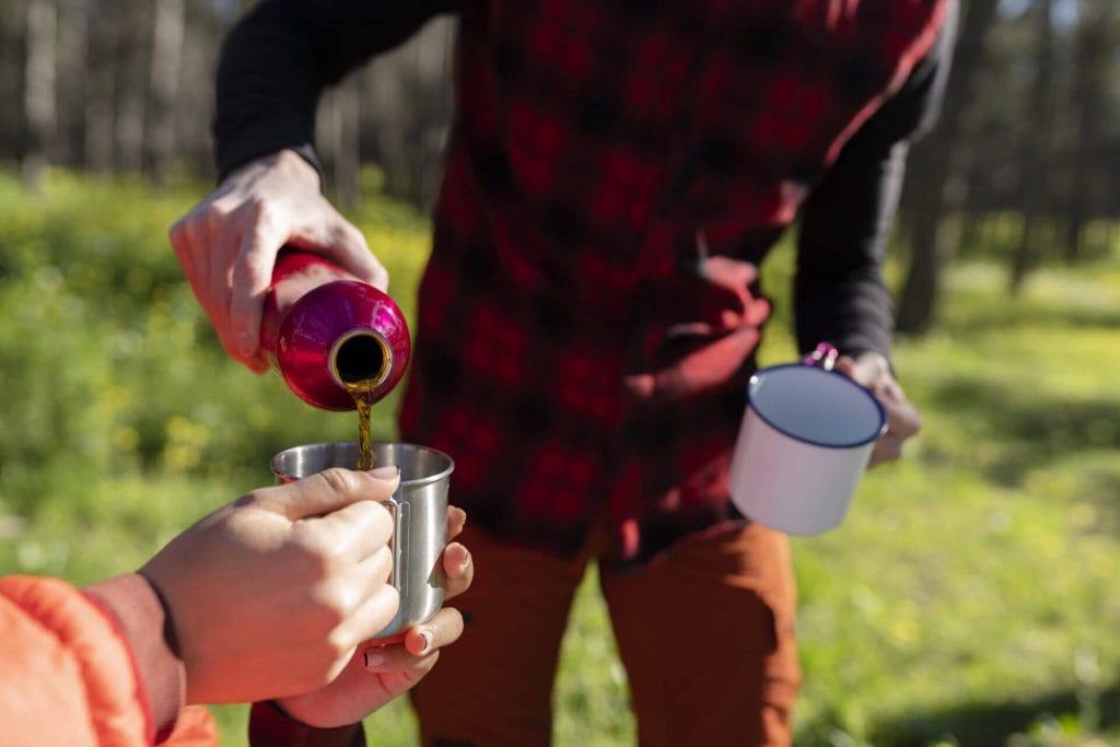 two adults camping, one pours another an adult beverage into a mug