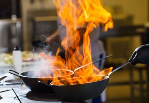 Close-up of a skillet on a stove with a tall flame, actively being shaken by the hands of a chef