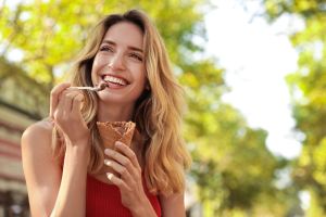 Woman standing outside and smiling, holding an ice cream cone in one hand, holding a spoon and scooping ice cream in the other hand