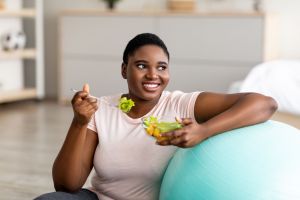 GLP-1 consumer sitting down, resting on an exercise ball, smiling and holding a salad and fork with salad on it, about to eat for her changed taste palate