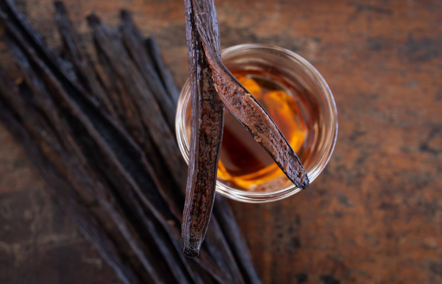 Vanilla bean pods and a glass of vanilla extract with two pods on top, all sitting on a table