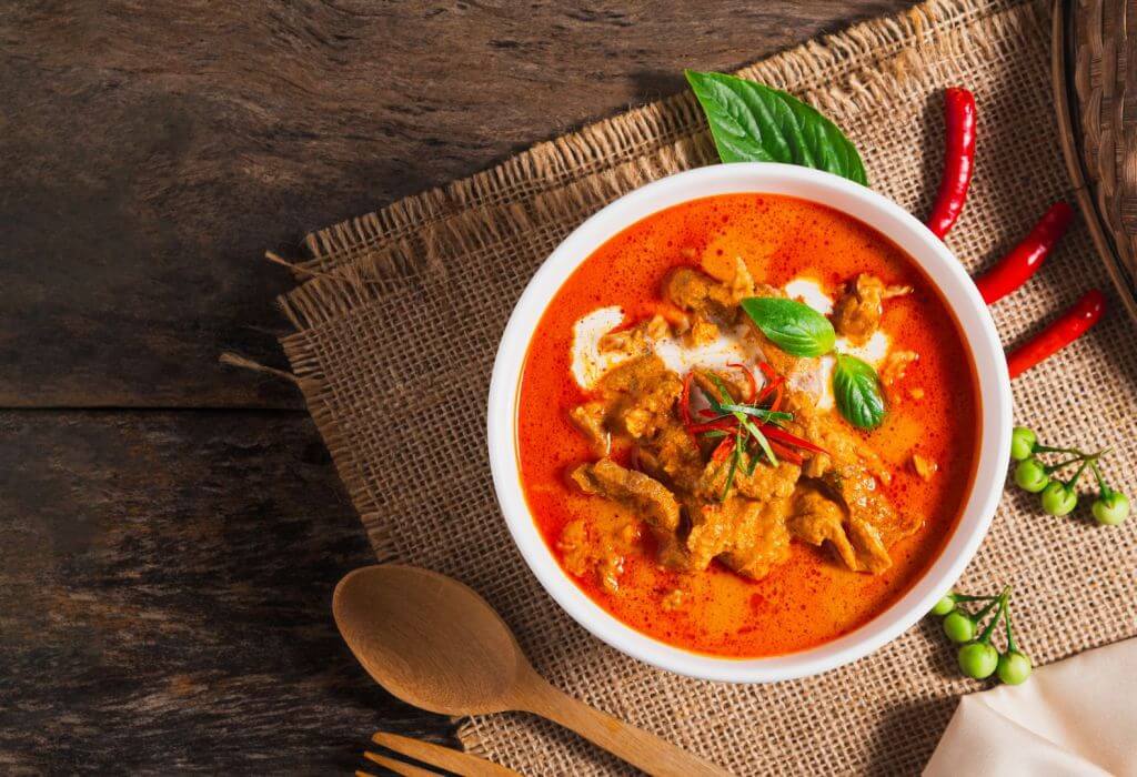 Red curry dish with meat and spices in a bowl, surrounded by chili peppers and other spices, flatware, place mat.