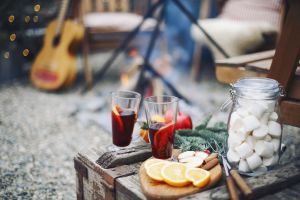 Glasses of non-alcohol mulled wine beverages sitting outside next to marshmallows and food on an outdoor table. In background, a campsite, guitar, outdoor fire pit and more